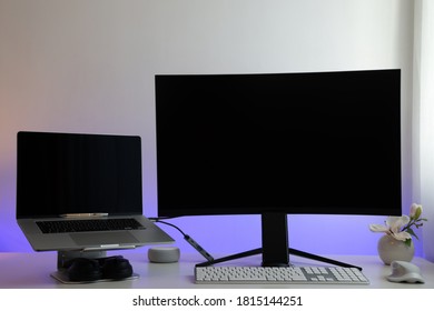 Modern Home Office Workplace With A Flat Laptop On A Stand, Black Headphones, A Large And Curved Monitor, A White German-language Keyboard, A White Mouse And A White Table