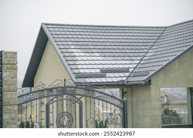 A modern home with a sleek gray roof, framed by an ornate metal gate, set against a cloudy sky. - Powered by Shutterstock