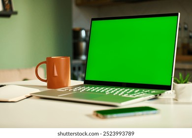 Modern home workspace featuring laptop with green screen, smartphone, coffee mug and small potted plants. Home office for freelancer with kitchen in background. Concept of productivity and remote work - Powered by Shutterstock