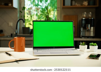 Modern home workspace featuring laptop with green screen, smartphone, coffee mug and small potted plants. Home office for freelancer with kitchen in background. Concept of productivity and remote work - Powered by Shutterstock