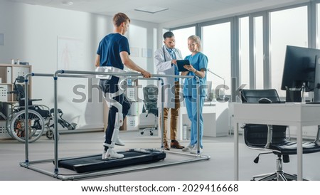Modern Hospital Physical Therapy: Patient with Injury Walks on Treadmill Wearing Advanced Robotic Exoskeleton Legs. Physiotherapy Rehabilitation Scientists, Engineers, Doctors use Tablet Computer