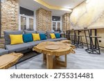 Modern hostel common area featuring large live-edge wooden tables, a grey sofa with yellow and blue pillows, and exposed brick walls. Includes a high wooden bar counter with stools and windows decorated with fairy lights. Trendy loft-style interior.