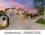 Modern house exterior with illuminated facade and landscaped front yard at sunset, showcasing a vibrant sky.