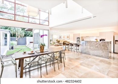 Modern House Interior And Outdoor Including Dining And Kitchen Beside Living Room, The Dining Or Patio Area Has A Wooden Table With Plastic See Through Chairs On The Tile Floor,  The House Illuminated