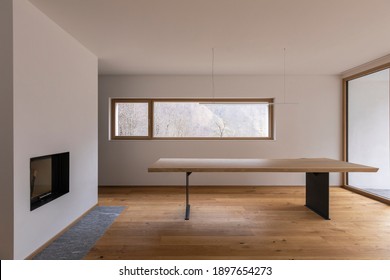 Modernes Interieur mit weißen Wänden und Parkettböden. Holztisch mit Designlampe, Kamin und Fenster zum Wald der Schweizer Alpen.  Niemand drinnen