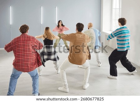 Modern indoor dance studio class, diverse group of dancing young people actively following trainer woman learning dance steps, team moving practicing choreography, feeling energized, rear view 