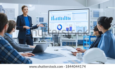 Modern Industrial Factory Meeting: Confident Female Engineer Uses Interactive Whiteboard, Makes Report to a Group of Engineers, Managers Talks and Shows Statistics, Growth and Analysis Information