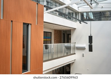 Modern interior architectural space featuring a multi-level atrium with clean, minimalist design. The left foreground shows a wall with vertical wood paneling and a window. The central area reveals multiple floors connected by glass balustrades with metal handrails. The right side features white walls with suspended black audio speakers hanging from the ceiling structure. The space includes track lighting fixtures, a wall-mounted air conditioning unit, and a glass roof or skylight allowing natural light to illuminate the area. In the background, exposed brickwork is visible on an upper level wall. A partial view of one person can be seen on the ground floor. The overall aesthetic combines wood, glass, and white surfaces creating a contemporary corporate or institutional environment.