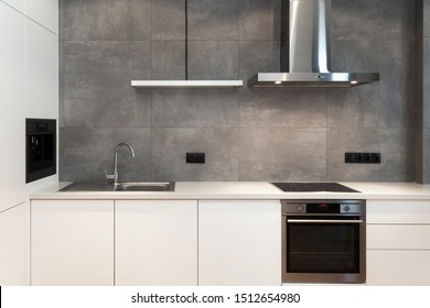 Modern interior design at white contemporary kitchen in loft style. Glossy cabinet with built in household appliance, electric stove, oven, sink on worktop and extractor hood on grey wall