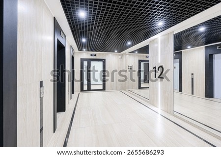 Modern interior with elevators, doors, and reflective surfaces. The ceiling is black grid, and the floor is tiled. Walls feature beige marble