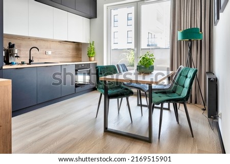 Modern interior of kitchen with dark furniture, dining table and stylish green chairs. Dining room with window and wooden floor. Stylish real estate.