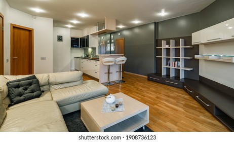 Modern Interior Of Kitchen In Luxury Studio Apartment. New Kitchen Set. Bar Stools. Cozy Leather Couch And Coffee Table. Wooden Doors. Bookshelf.