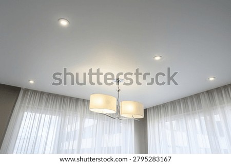 Modern interior with a matte white stretch ceiling, recessed LED spotlights, a contemporary chandelier with cream shades, and sheer white curtains framing the windows