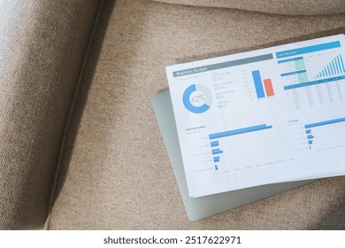 A modern interior with soft lighting, featuring a sofa, glass-walled room, and a shelf. A laptop and financial chart on A4 paper rest on the carpet, creating a productive yet calm space.