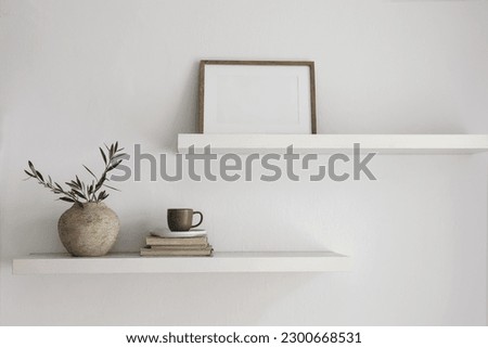 Modern interior still life. Two floating shelves. Blank wooden picture frame mockup template. Vase with olive tree branches, old books, cup of coffee. Modern Mediterranean home. White wall background.