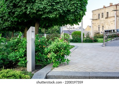 modern iron lantern of ground garden led lighting with bushes in city street with trees near flower bed landscape next to the ramp and steel railings, in the background is an old town house, nobody.