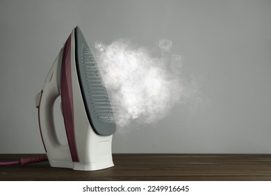 Modern iron with steam on wooden table against light grey background, space for text