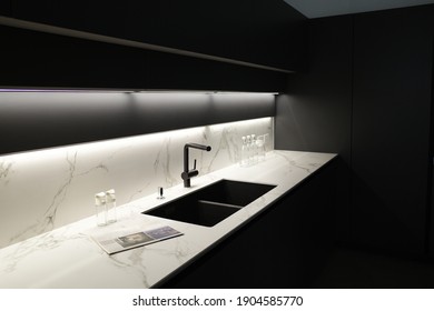 Modern kitchen with black furniture, White marble worktop and backsplash.Black sink and tap, Light under wall cabinet.