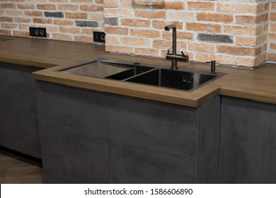 Modern Kitchen With Black Sink, Kitchen Faucet For Water And Fronts Against The Background Of A Brick Wal. Brown And Black Wooden Countertops. Luxury Dark Themed Background For Interior.