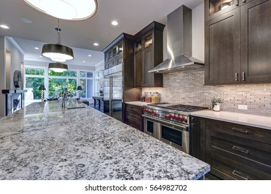 Modern kitchen with brown kitchen cabinets, oversized kitchen island, granite countertops, stainless steel hood over six burner Range and beige backsplash. Northwest, USA
