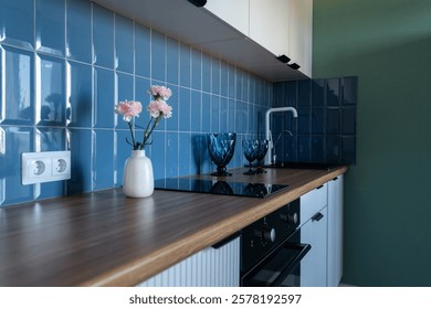 Modern kitchen with cabinets and induction on counter. minimalist kitchen design with white cabinets, blue backsplash, wooden countertop. vase of pink flowers and blue glasses on table top. 
