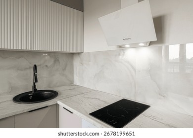 A modern kitchen corner features light cabinets, a black sink, a white range hood, and marble backsplash. A sleek black cooktop sits on the counter