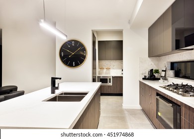 Modern Kitchen Counter Top Closeup With A Stove And Watch Beside The Oven And Pantry Cupboards, The Counter Included A Tap And Sink Under The  Hanging Tube Light