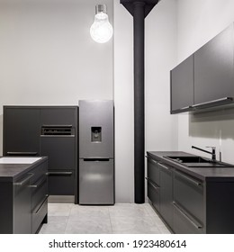 Modern Kitchen With Dark Gray Cupboards, Drawers And Countertops, Silver Fridge And Gray Floor Tiles And Decorative Black Pillar And Light Bulb In Loft Apartment