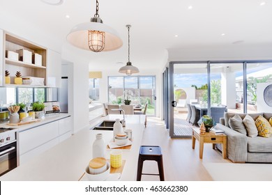 Modern Kitchen With A Dining And Patio Area Including Some Utensils On Counter Top Under The Hanging Lamps Closely, The Living Room With Sofa And Pillows Beside Outdoor Patio Area With Sunlight 