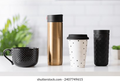 Modern Kitchen Drinkware Assortment on a White Countertop: Textured Black Ceramic Mug, Shiny Gold Insulated Flask, White Plastic Cup with Black Dots, and a Matte Black Tumbler with a Geometric Pattern - Powered by Shutterstock