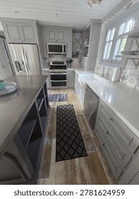 A modern kitchen interior featuring light grey cabinetry white stone countertops and stainless steel appliances. The space includes a central island and rustic wooden flooring.