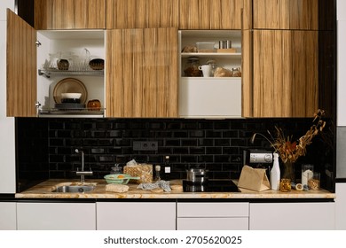 Modern kitchen interior featuring open wooden cabinets displaying plates, glasses, and jars, countertop holding pasta, bread, coffee machine, pot, and dried flowers, black tile backsplash