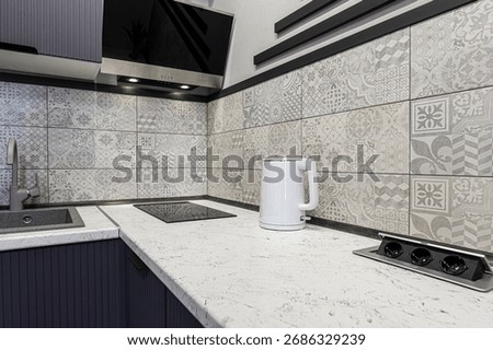 Modern kitchen with patterned backsplash, white countertop, and a white electric kettle. Features a sleek cooktop and a hidden power outlet on the counter