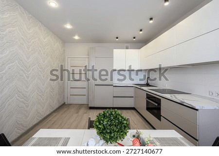 modern kitchen with white upper cabinets, beige lower units, marble-like countertops, textured wallpaper, recessed ceiling lights, dining table with greenery centerpiece