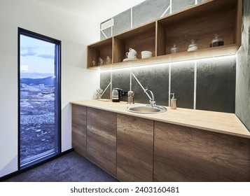 Modern kitchen with white walls and glass door with scenic view inside. Side view of kitchen area with wooden shelves and working surface, illuminated with warm LED light indoor. Concept of interior.