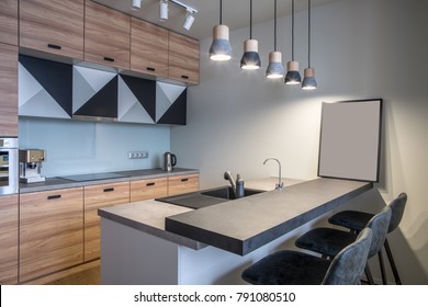 Modern Kitchen With White Walls And Luminous Hanging Lamps. There Is A Light Kitchen Island With A Sink And Gray Tabletops And Dark Chairs, Wooden Lockers, Oven, Stove, Chrome Kettle, Coffee Maker.