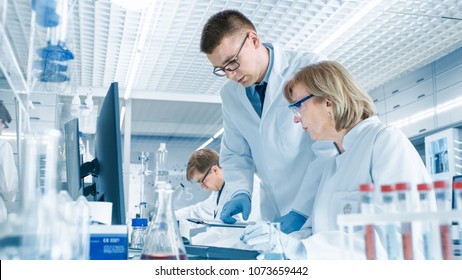 In Modern Laboratory Senior Female Scientist Has Discussion with Young Male Laboratory Assistant. He Shows Her Data Charts on a Clipboard, She Analyzes it and Enters It into Her Computer.