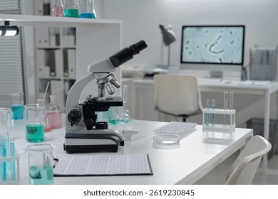 Modern laboratory setup featuring high-tech microscope and various chemical solutions on workbench. Background includes organized workspace with computer showing magnified cellular image