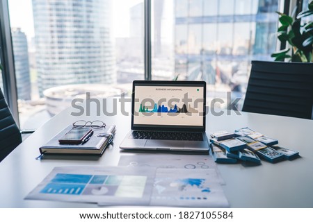 Modern laptop computer with infographic statistics for business displayed on screen staying on table desktop in office interior of corporate company, netbook and wat of money as concept of wealthy