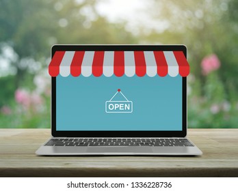 Modern Laptop Computer With Online Shopping Store Graphic And Open Sign On Wooden Table Over Blur Pink Flower And Tree In Garden, Business Internet Shop Online Concept