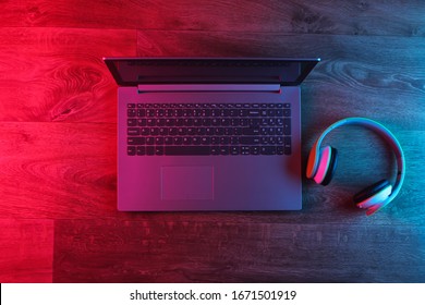 Modern Laptop With Headphones On A Wooden Floor. Blue Red Neon Light. Gradient Glow. Gaming Or DJ Still Life. Top View