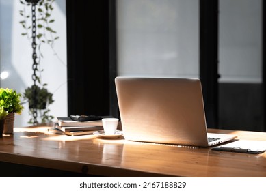 A modern laptop on a wooden desk in a cozy office setting with natural light, books, a plant, and a cup of coffee. - Powered by Shutterstock