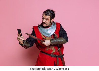 Modern Lifestyle. Comic Portrait Of Medieval Warrior Or Knight Wearing Wearing Armor Clothing Using Phone Isolated Over Pink Background. Comparison Of Eras, History, Funny Meme Emotions