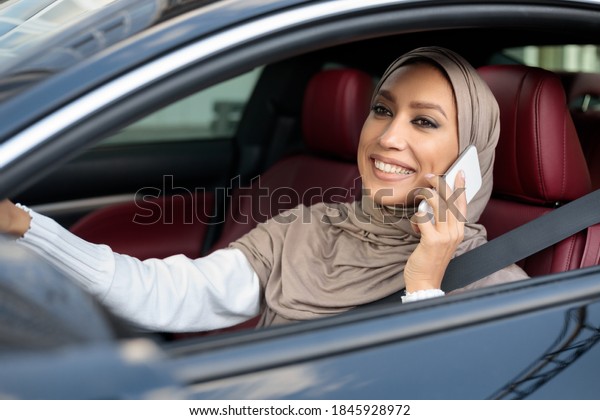 Modern Lifestyle. Portrait of smiling muslim woman\
in hijab driving new luxury car, talking on mobile phone. Cheerful\
arabian lady in headscarf sitting in salon on driver\' seat, holding\
steering wheel