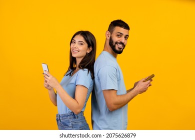 Modern lifestyle. Positive arab couple standing back to back, holding and using their mobile phones, chatting online or scrolling social networks feed, yellow studio background