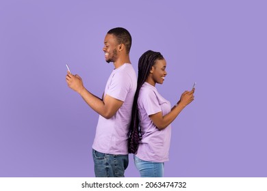 Modern Lifestyle. Profile side view portrait of cheerful positive African American couple standing back to back, holding and using their mobile phones and chatting online, violet studio background