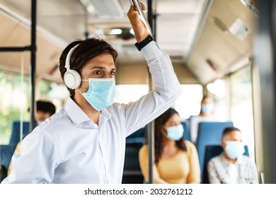 Modern Lifestyle. Young man wearing white shirt, surgical face mask and wireless headset traveling by tram in town, standing and looking out of window, listening to music, holding handle. Healthcare - Powered by Shutterstock