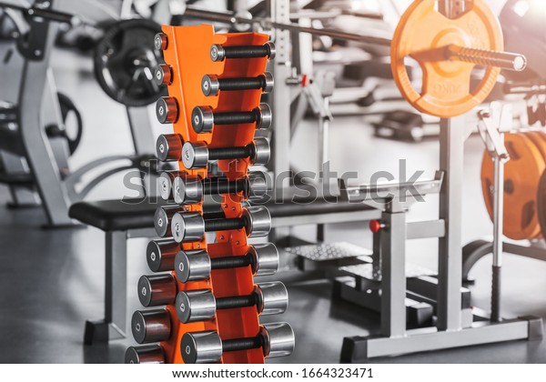 Modern light gym. Sports equipment in gym.\
Dumbbells of different weight on rack. Design of the room and\
equipment in gray and\
orange