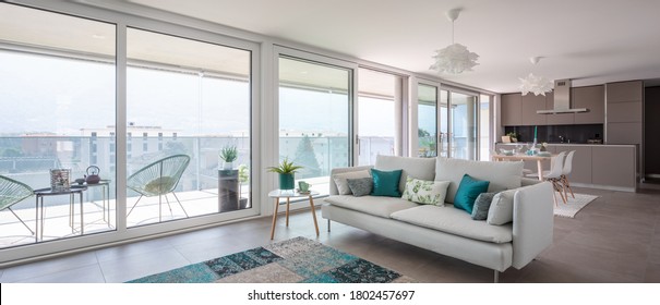 Modernes Wohnzimmer mit Designer-Möbeln. Sofa mit hellen Kissen und Balkon-Aussicht. Hinter der modernen Küche mit Insel. Niemand drinnen. 
