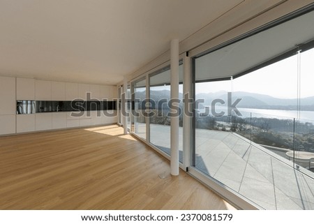Modern living room with open space and a black and white designed kitchen situated at the end. Large windows allow plenty of sunlight to enter. The room features a parquet floor. Nobody inside